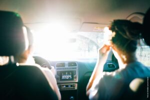 Zwei Personen sitzen im Auto und werden von hinten gezeigt. Von vorne strahlt Sonne in das Auto.