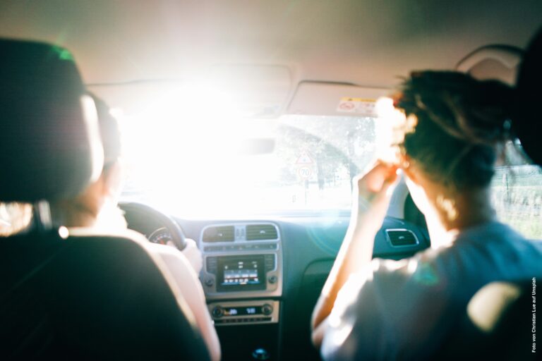 Zwei Personen sitzen im Auto und werden von hinten gezeigt. Von vorne strahlt Sonne in das Auto.